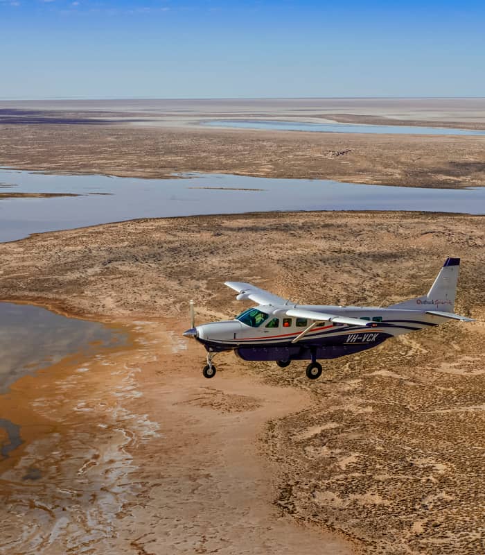 Aerial perspective of an Outback Spirit aircraft navigating the desert fringes of Lake Eyre, where the red earth of the Tirari Desert meets the filling salt pans.