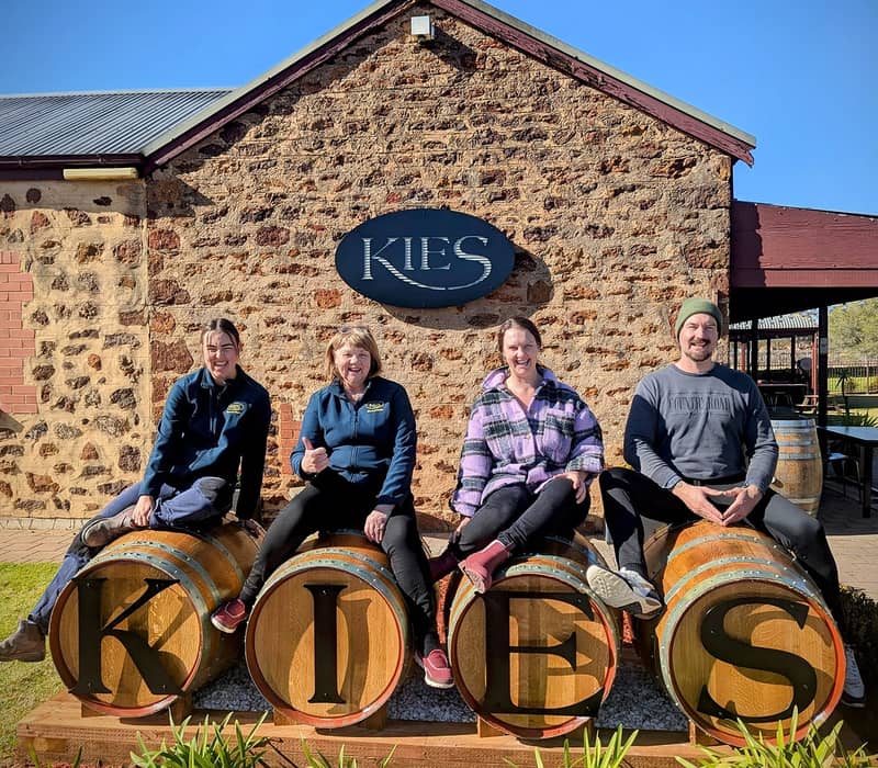 Four people sitting on large wine barrels that spell out KIES in front of a rustic stone winery building.