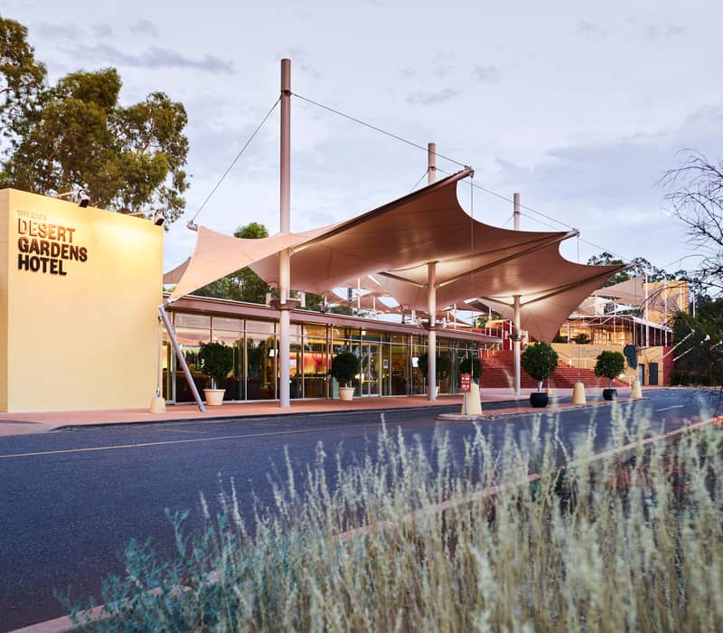 Luxury hotel room with a king bed and glass doors leading to a private balcony with a direct view of Uluru in the distance.