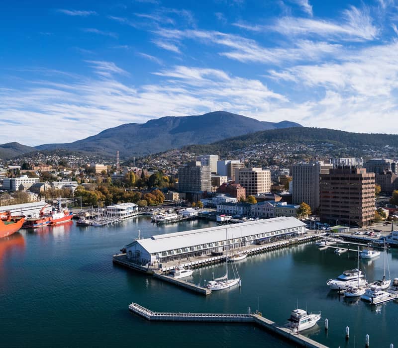 Bright harbor view of Hobart with ships, sailboats, and city buildings under a wispy blue sky.