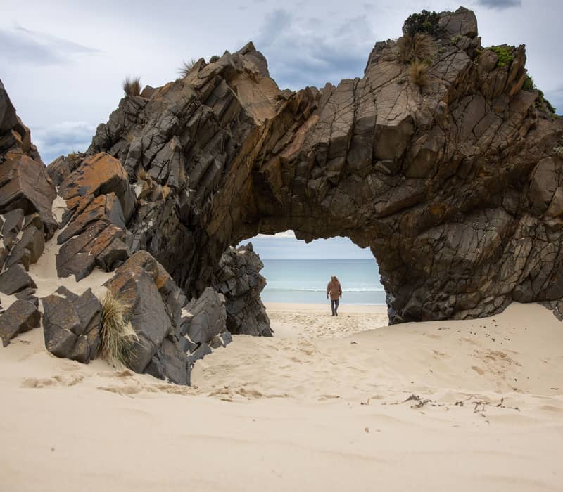 A natural rocky archway stands on a sandy beach at Mars Bluff, with a hiking trail winding through coastal heath and views of the blue ocean in the background.