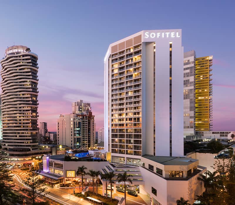 A sparkling blue swimming pool surrounded by white lounge cabanas and high-rise buildings at sunset.