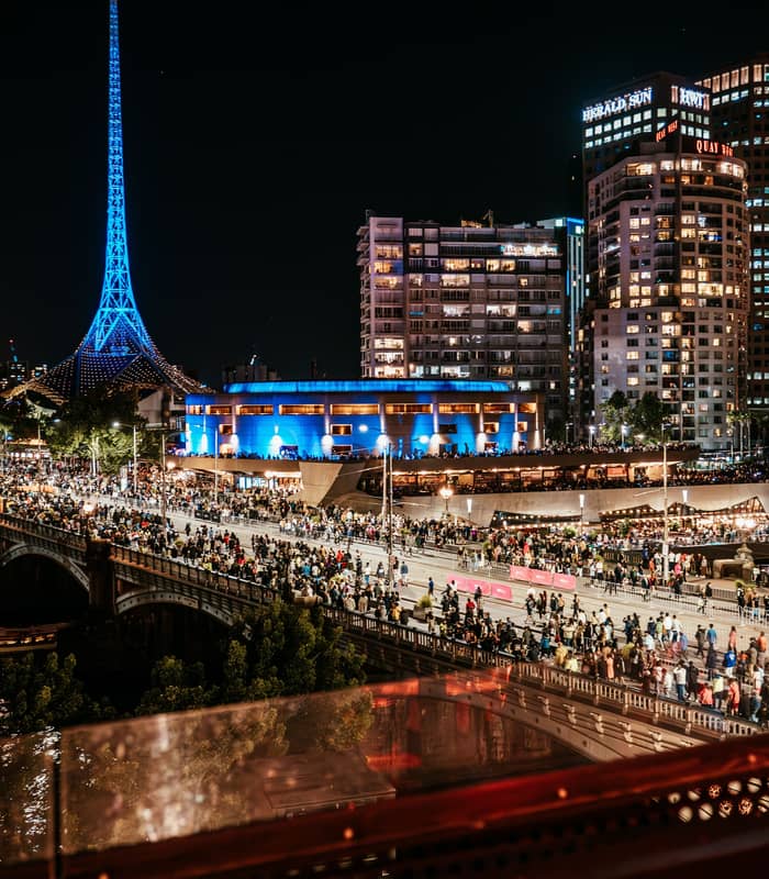 night scene looking down from a balcony over a busy bridge and river towards a tall illuminated blue spire.