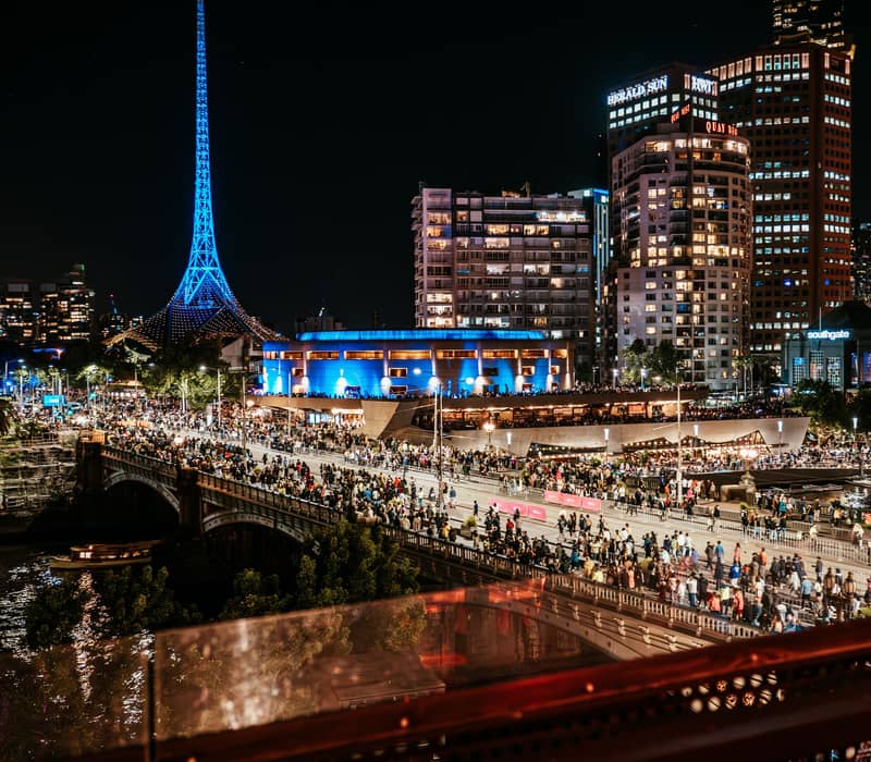 night scene looking down from a balcony over a busy bridge and river towards a tall illuminated blue spire.