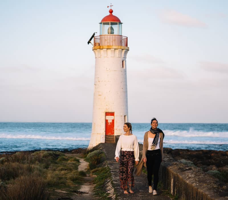 Two women walking along a stone causeway toward a white lighthouse with a red top at Port Fairy.