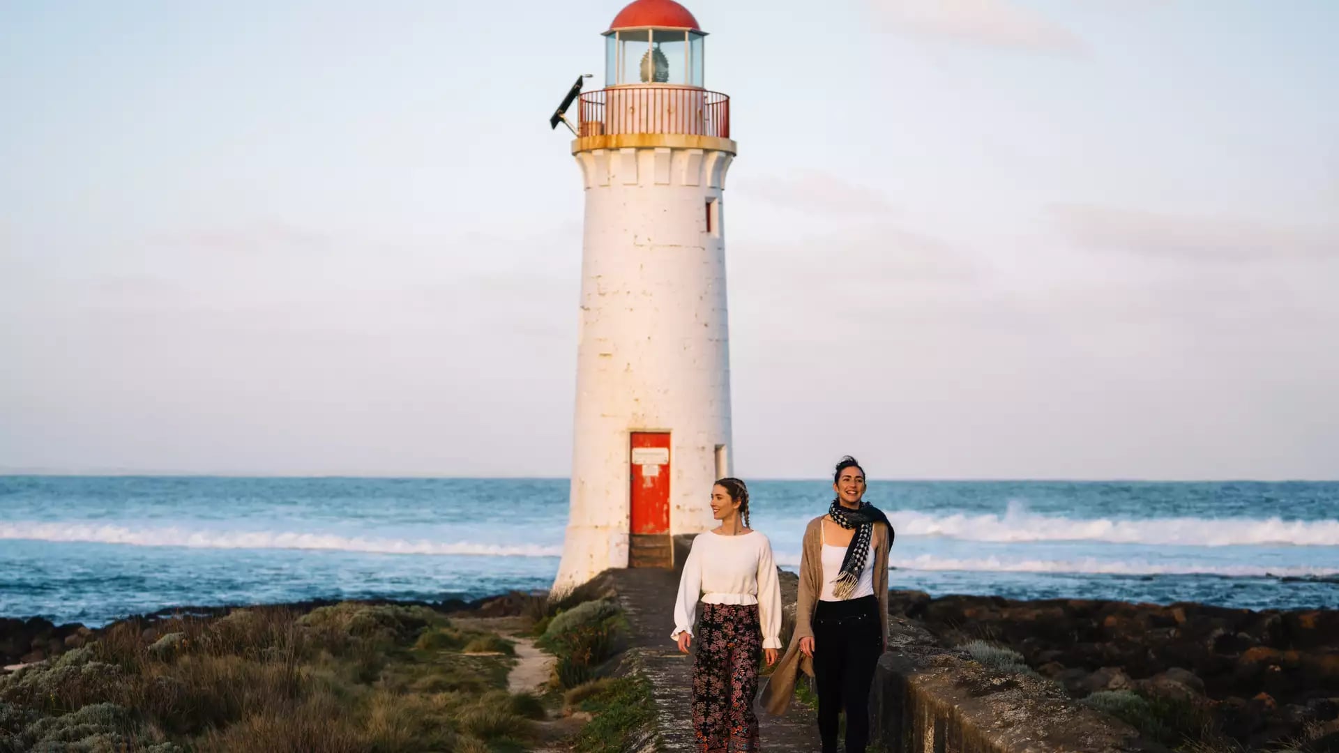 Two women walking along a stone causeway toward a white lighthouse with a red top at Port Fairy.