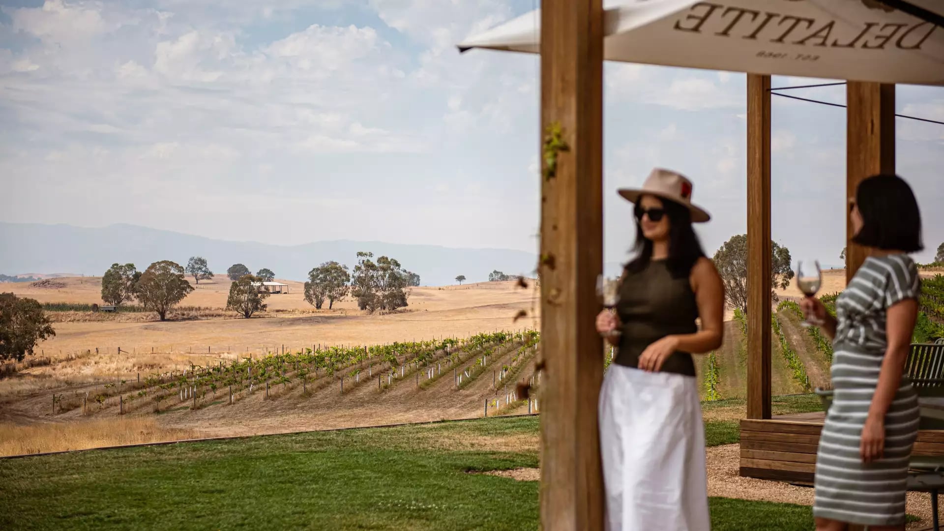 Two women stand on a timber deck drinking white wine, with a view of green vineyards and rolling yellow hills.