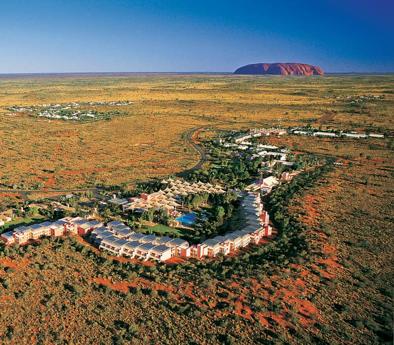 High-angle aerial view of the desert resort buildings and swimming pools with Uluru visible in the far distance under a clear blue sky.