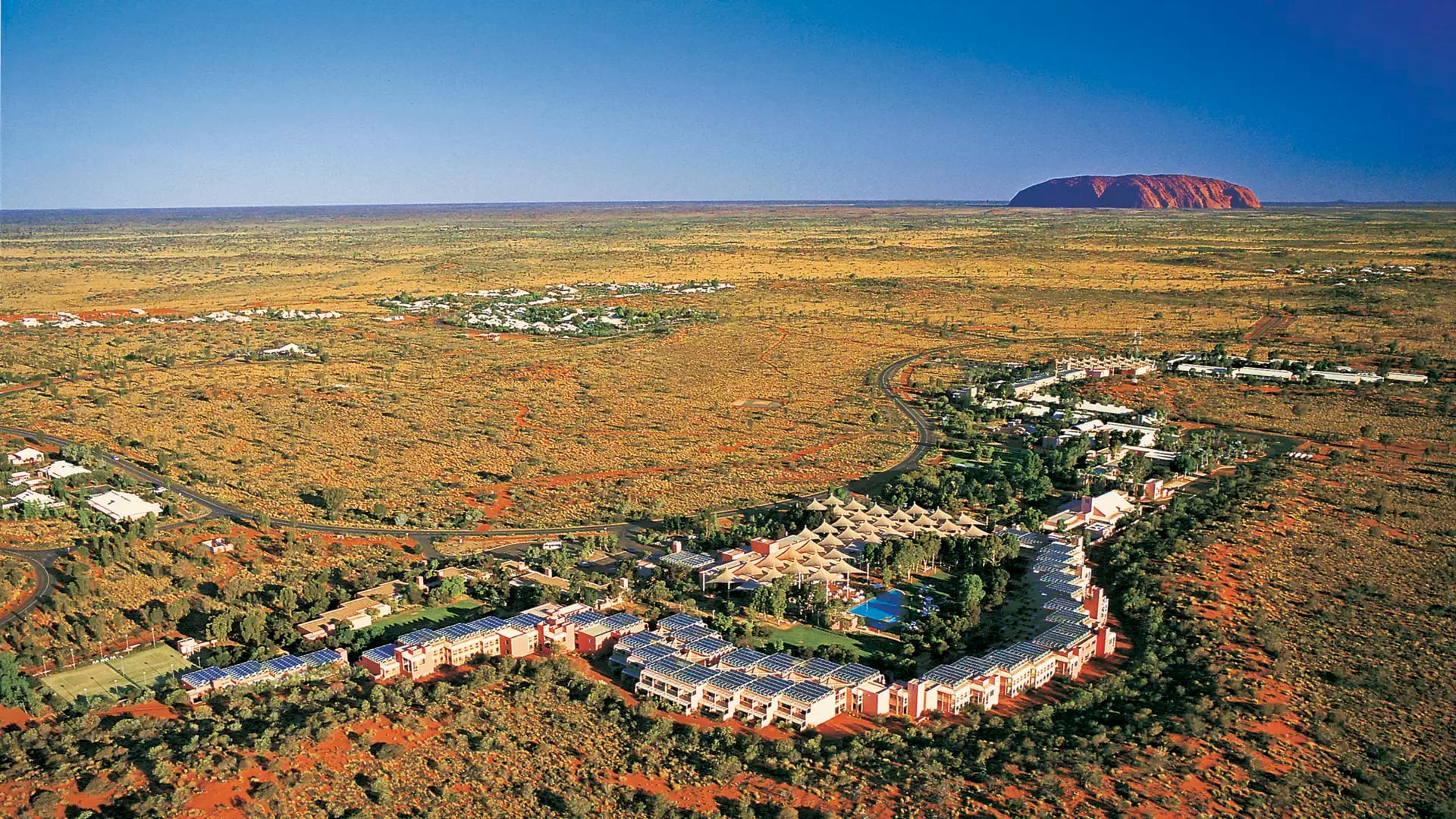 High-angle aerial view of the desert resort buildings and swimming pools with Uluru visible in the far distance under a clear blue sky.