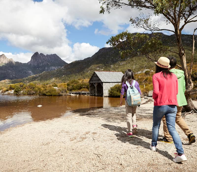 A group of tourists of three walking along the pebbled shore of Dove Lake towards the iconic boat shed at Cradle Mountain, Tasmania.