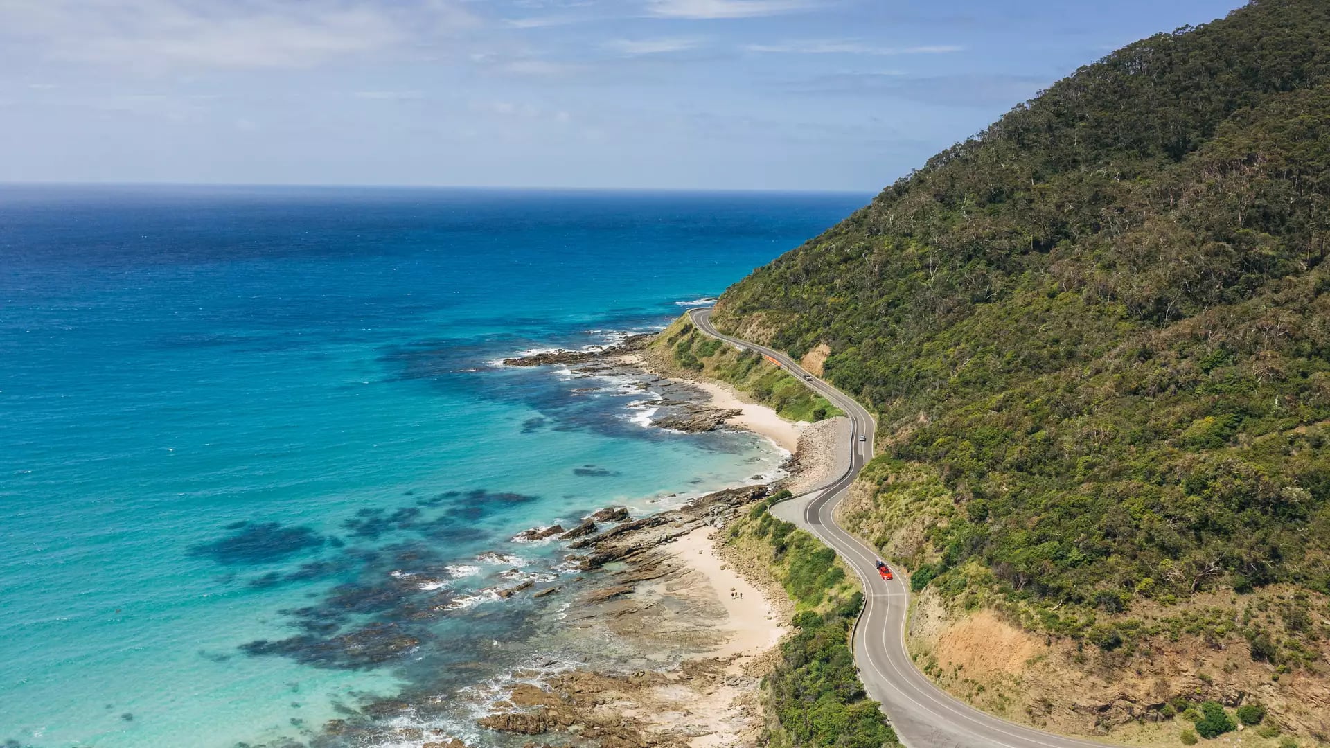 High-angle view of the Great Ocean Road winding along a turquoise coastline and green hills.