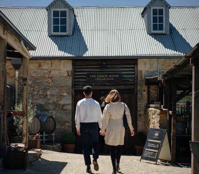 A man and woman holding hands as they walk toward the historic sandstone 'The Coach House Cellar Door' building in Richmond.