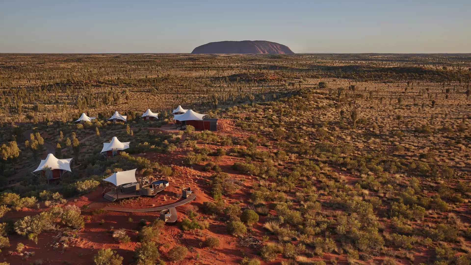 High-angle shot of sixteen white peaked-roof tents arranged in a curve along bright orange sand dunes, with sparse green desert oaks and the large red rock of Uluru on the horizon.