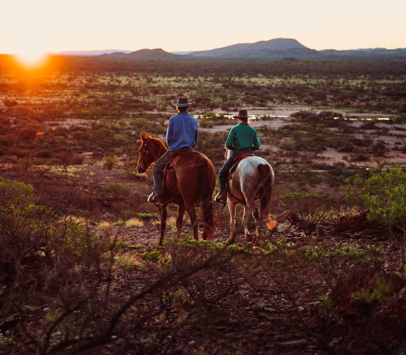 Two people riding horses through the Australian outback during a vibrant orange sunset in the Pilbara.