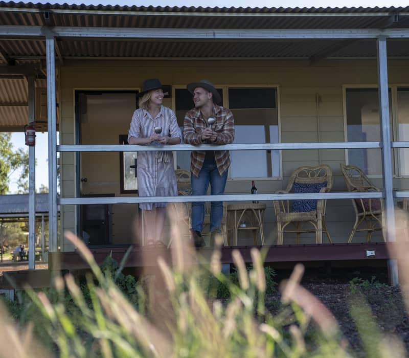 A man and woman in outback hats leaning on a porch railing with wine glasses at an Australian station stay.