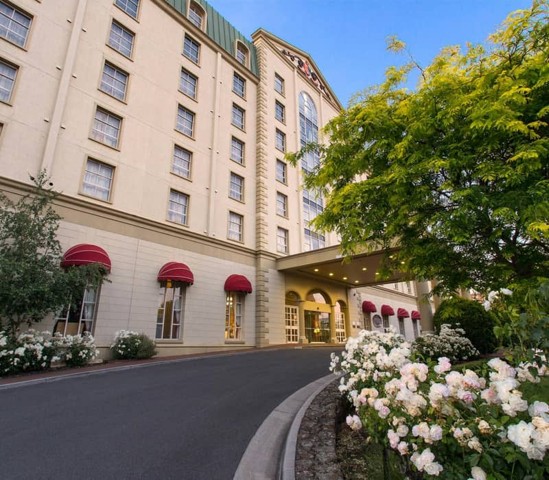Hotel entrance with driveway, white flowers, green trees, and beige building with red awnings.
