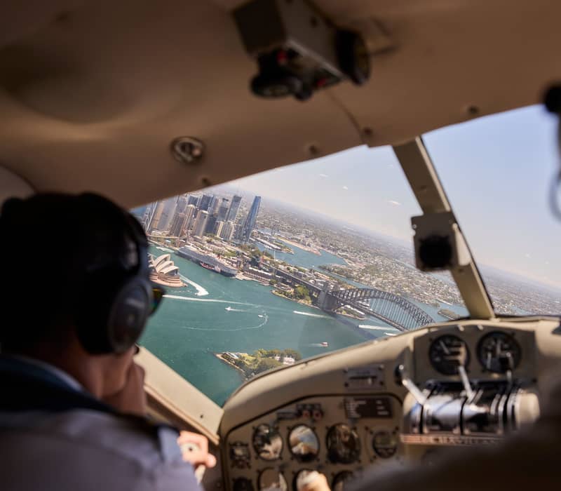 Cockpit view of a seaplane flying over the Sydney Harbour Bridge and city skyline.