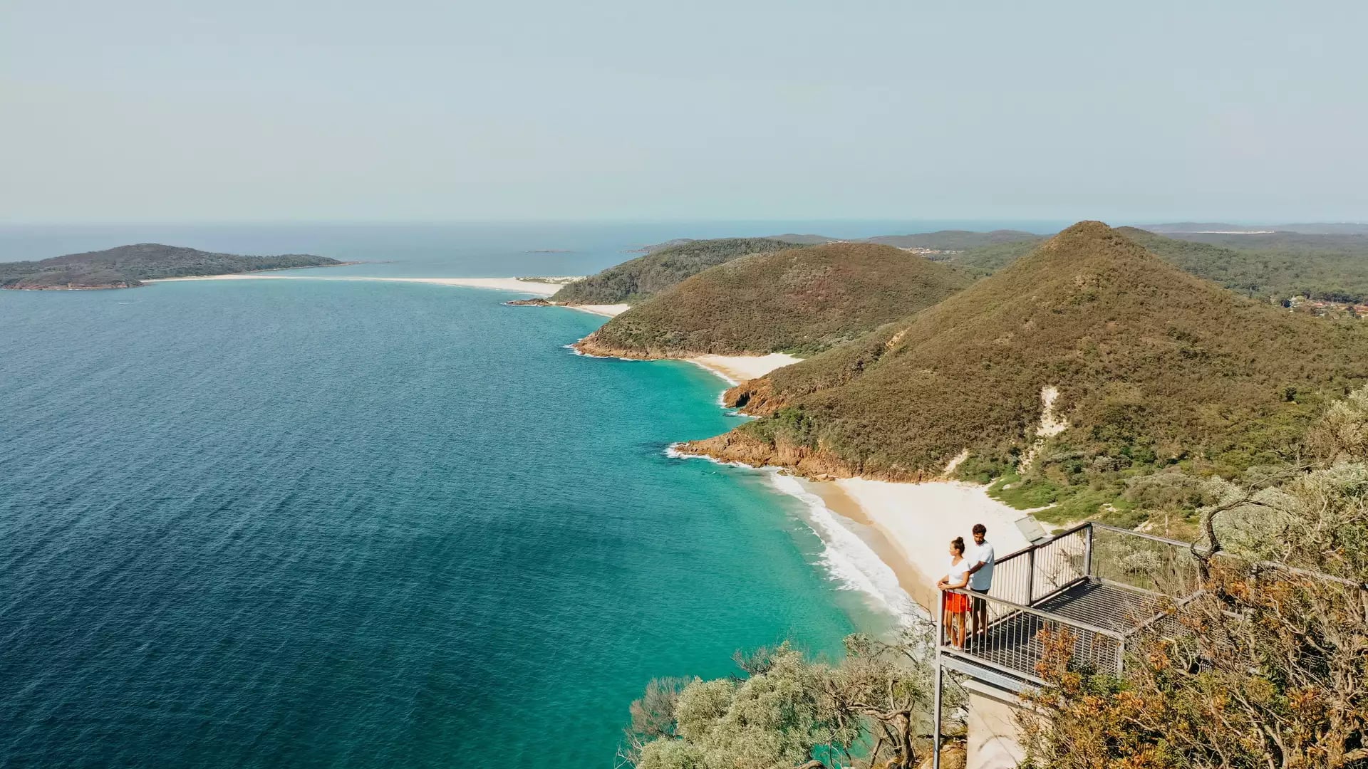 Couple standing on a metal lookout platform overlooking a turquoise bay and green coastal mountains.