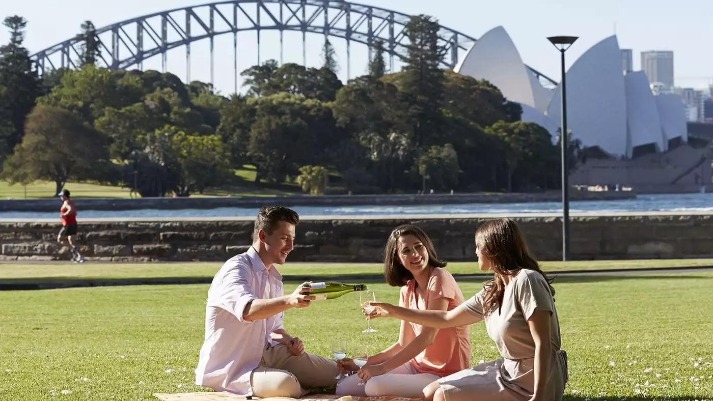 Three friends sitting on a picnic blanket in a park with the Sydney Opera House and Harbour Bridge visible behind them.