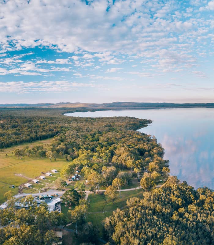 An expansive aerial panorama of the green camping grounds at Habitat Noosa bordering the calm, reflective waters of the Noosa Everglades in Queensland.