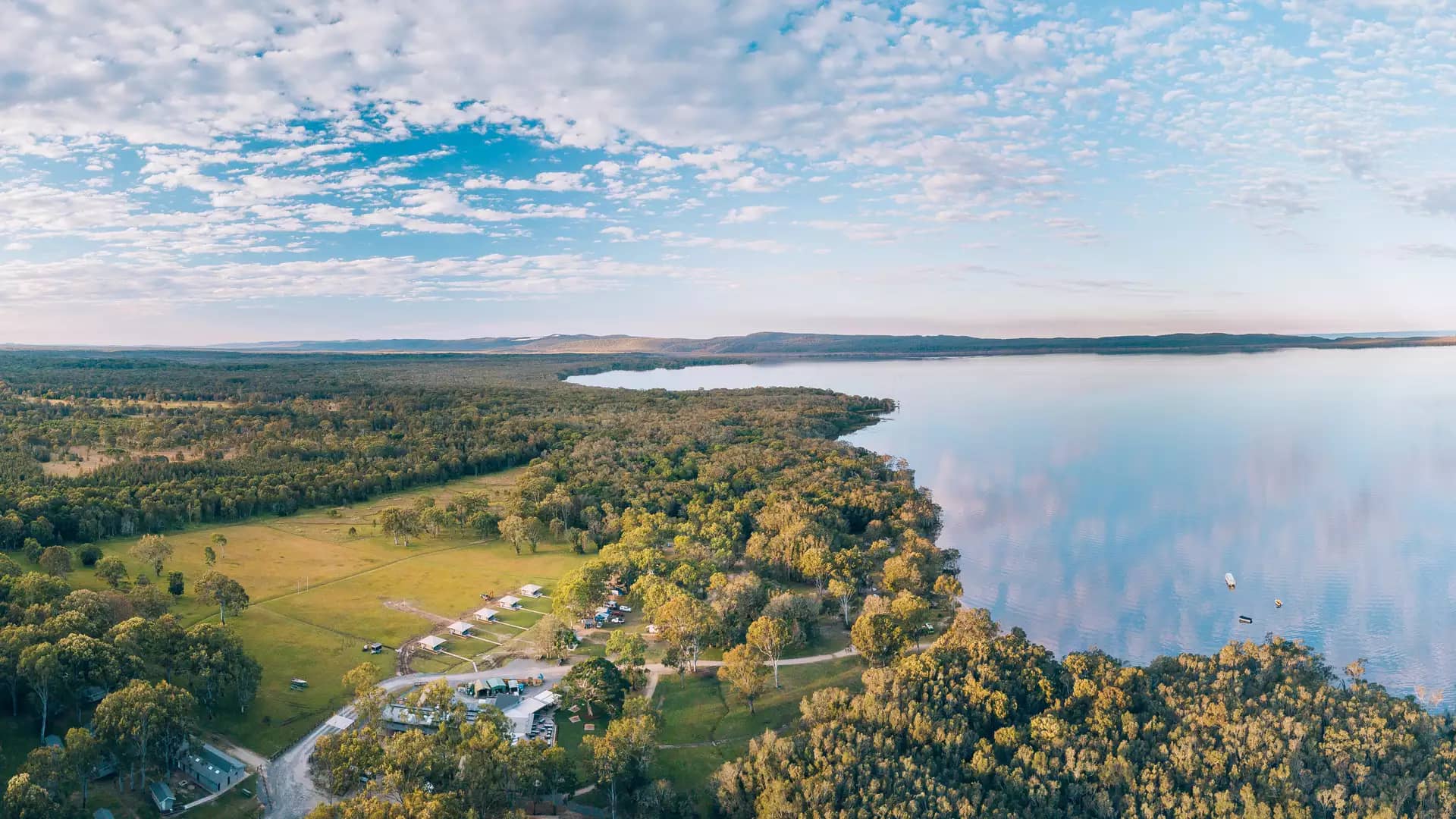 An expansive aerial panorama of the green camping grounds at Habitat Noosa bordering the calm, reflective waters of the Noosa Everglades in Queensland.