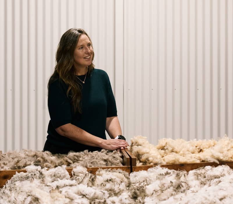 A woman with long brown hair, wearing a dark top, sits behind a large pile of raw, light-colored wool, looking off to the side and smiling slightly
