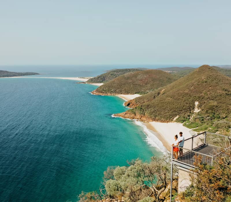 Couple standing on a metal lookout platform overlooking a turquoise bay and green coastal mountains.