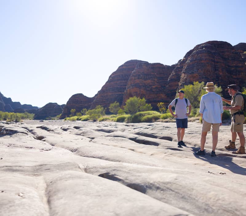 Three people standing on a textured rock surface looking toward high sandstone mountains in the Kimberley outback.