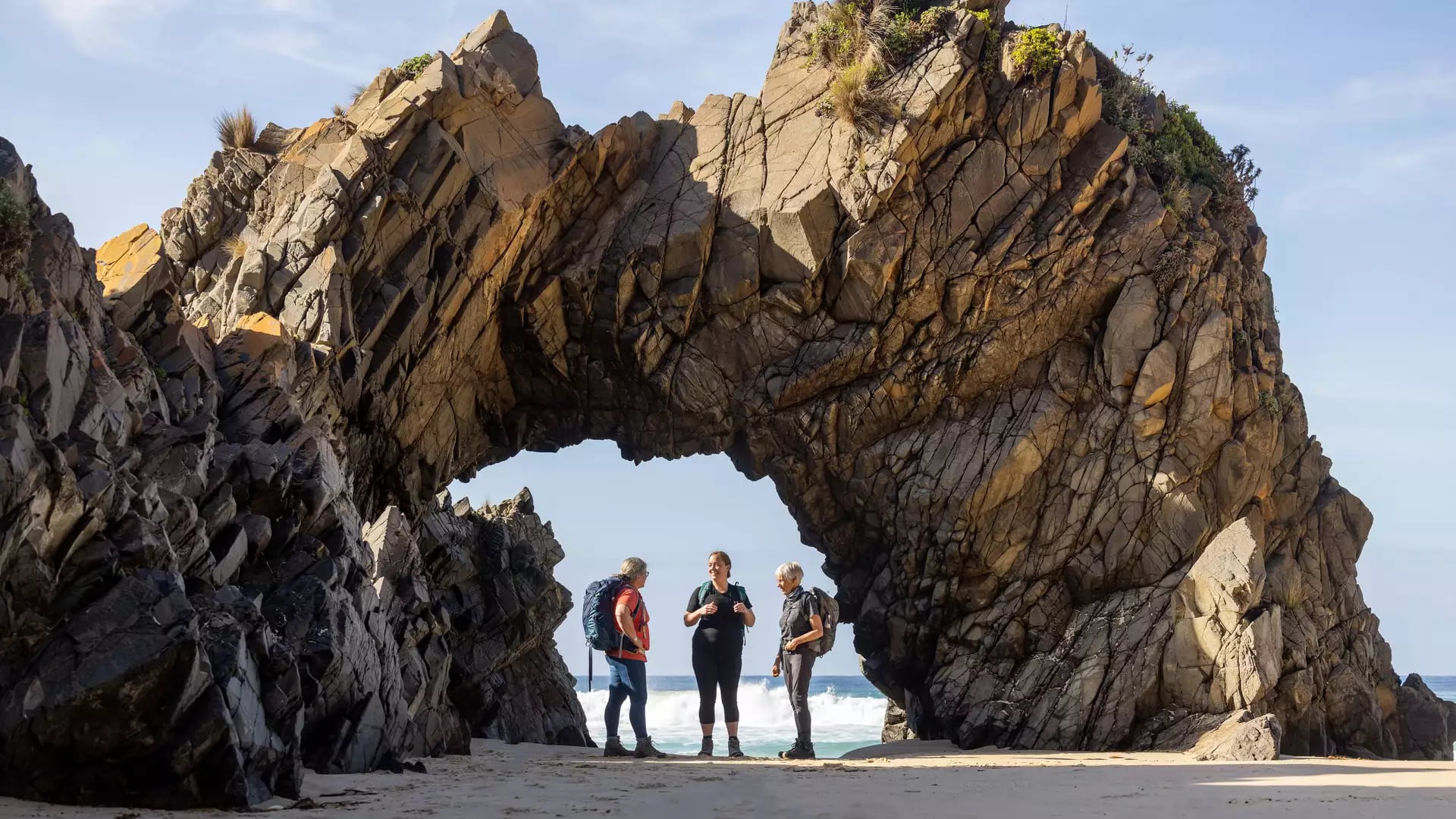 Travelers standing under a large natural rock formation arch on a beach in Tasmania.