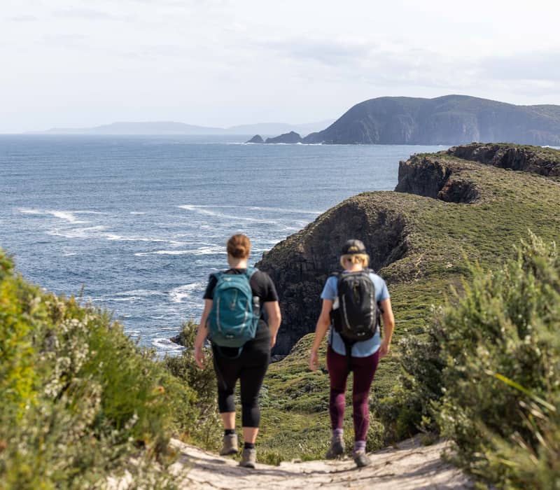Two hikers with backpacks walking toward a dramatic coastal cliff overlooking the ocean in Tasmania.