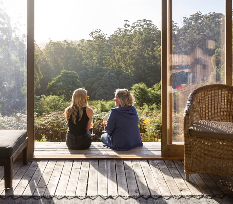 Two women sitting on a wooden deck overlooking a lush green forest at sunset in Tasmania.