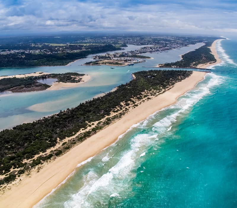 High-angle drone shot of a long thin peninsula with green trees and white sand beaches between blue lake water and the ocean.
