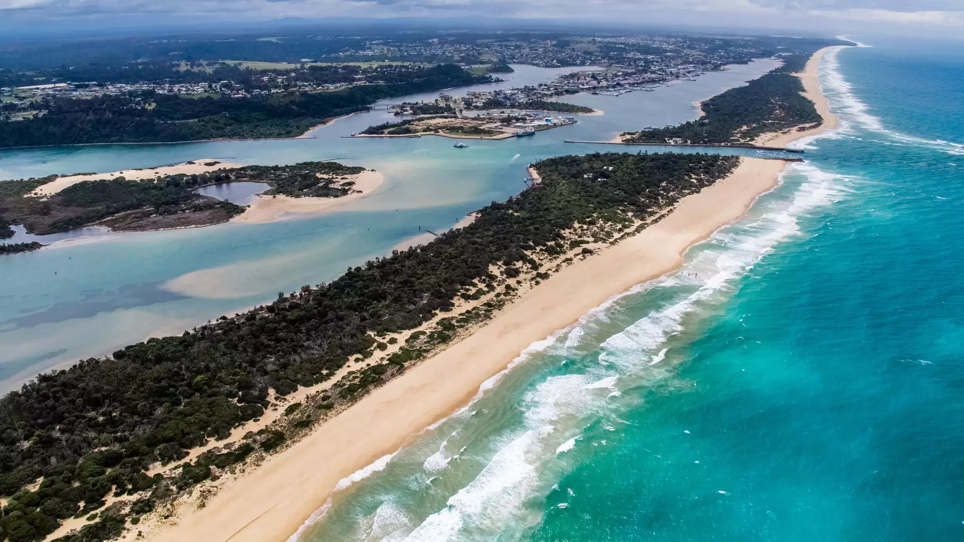 High-angle drone shot of a long thin peninsula with green trees and white sand beaches between blue lake water and the ocean.