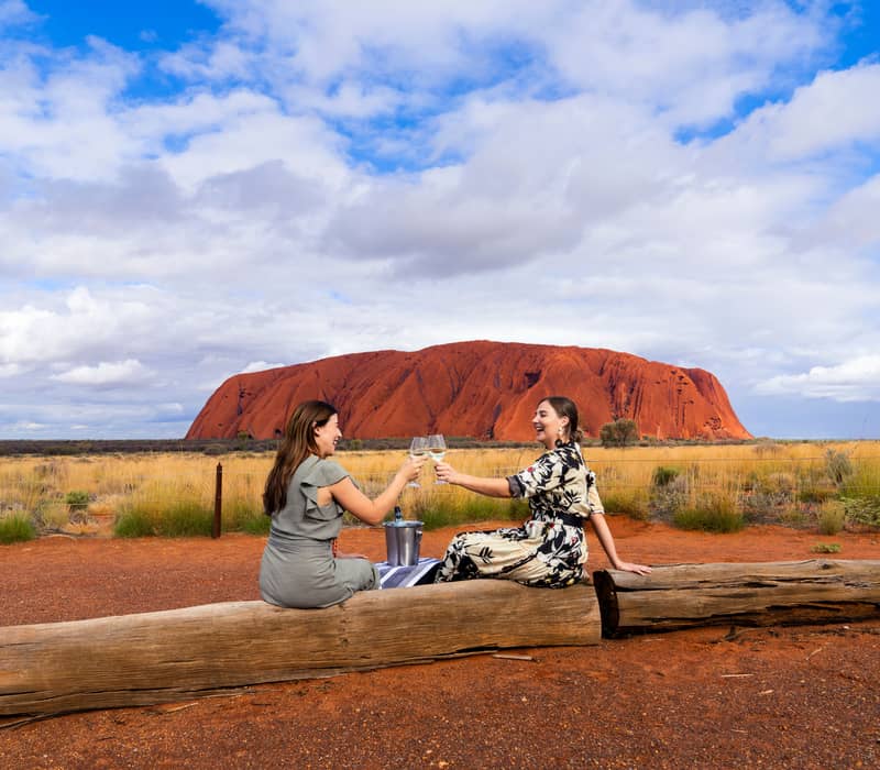 Two women toasting with wine glasses at sunset in front of Uluru, Northern Territory, Australia.