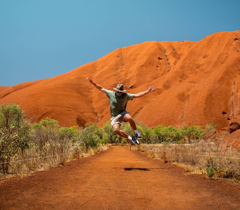 A man jumping for joy on a hiking path at the base of Uluru, Northern Territory, Australia.