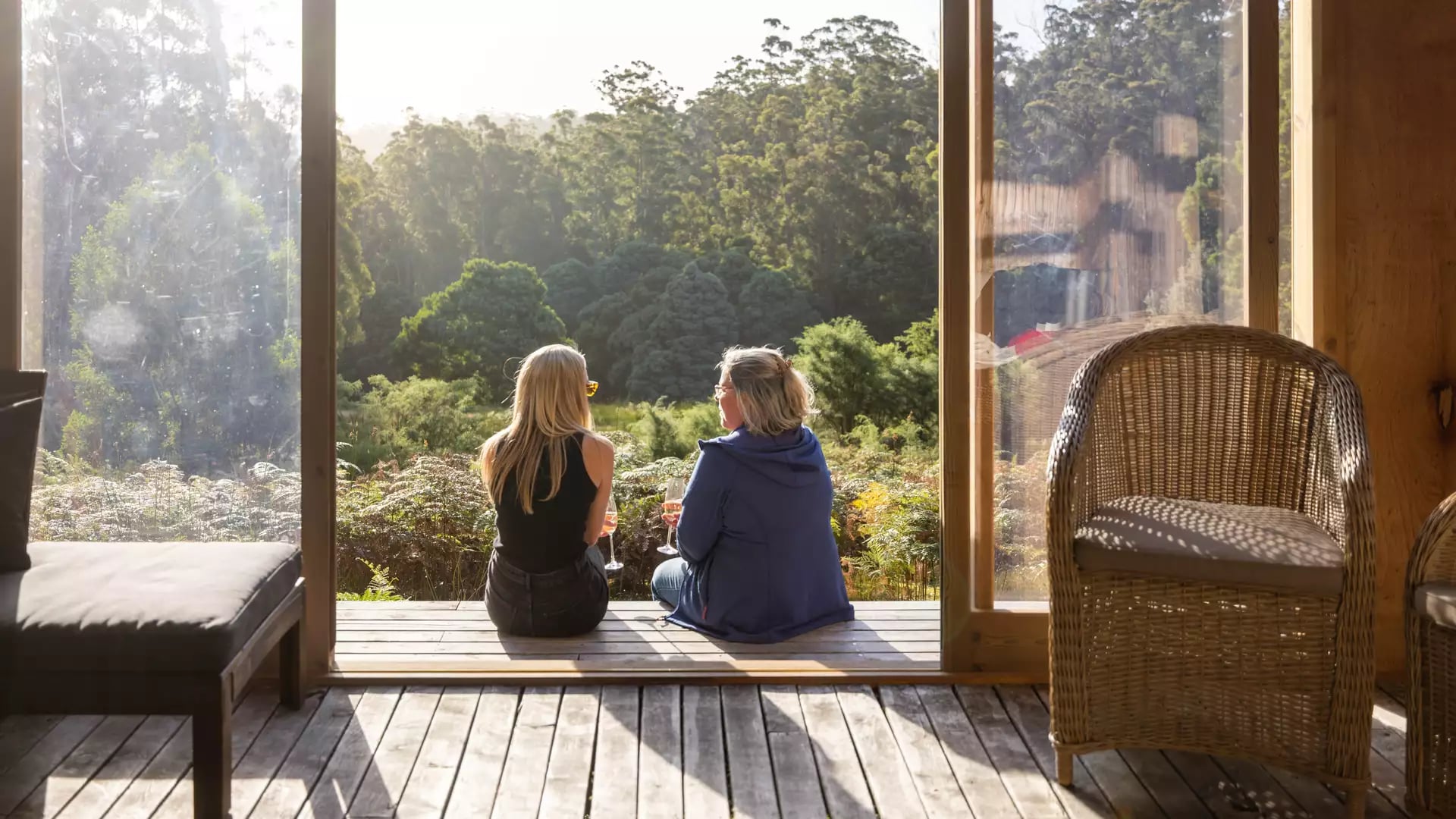 Two women sitting on a wooden deck overlooking a lush green forest at sunset in Tasmania.