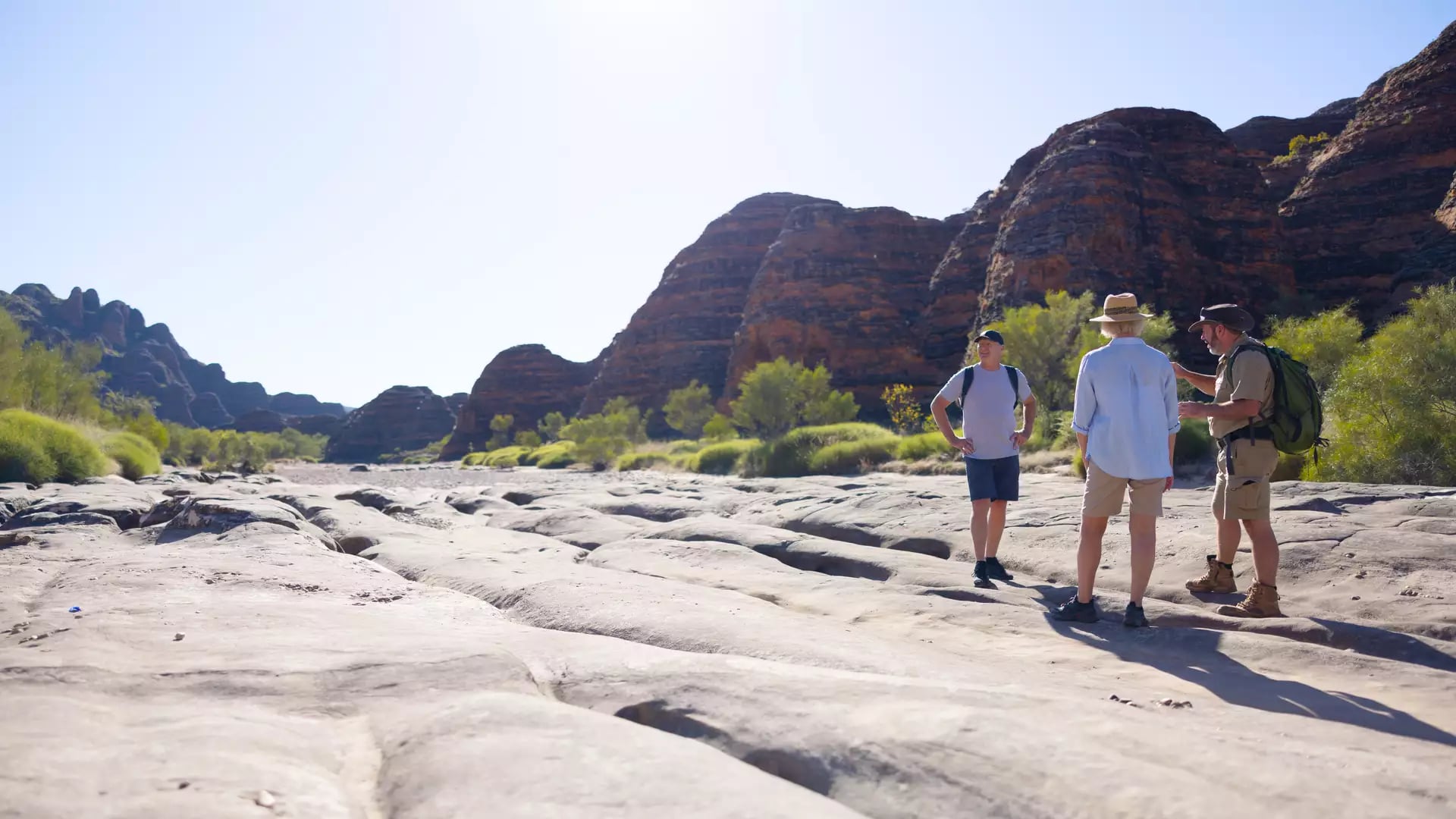 Three people standing on a textured rock surface looking toward high sandstone mountains in the Kimberley outback.
