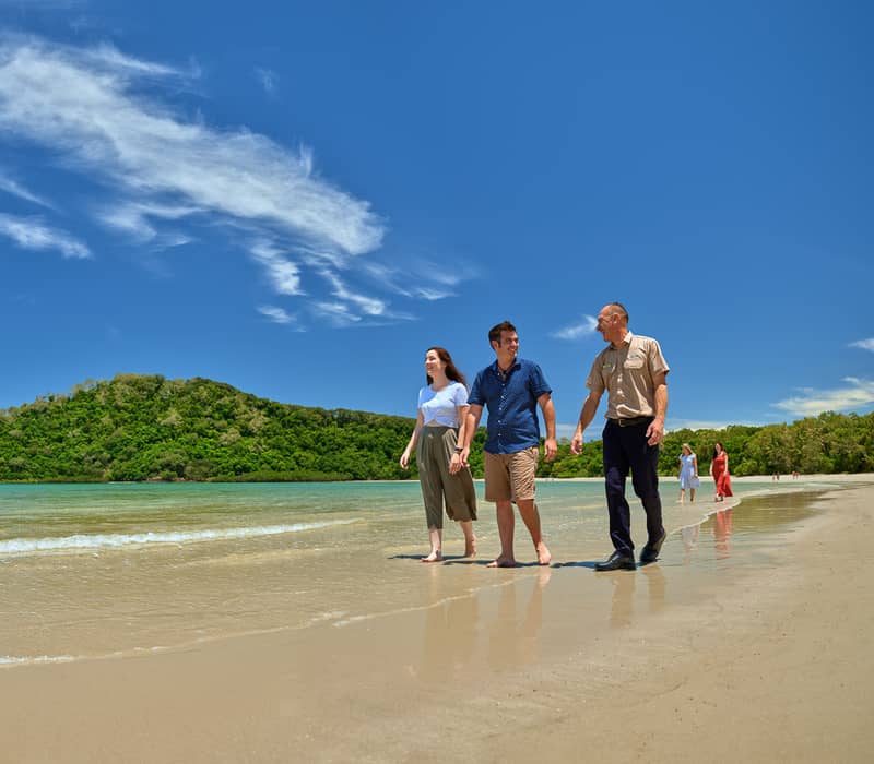 A small group of tourists walking along a wide, shallow sandy beach with a local Kuku Yalanji guide under a bright blue sky with wispy clouds.