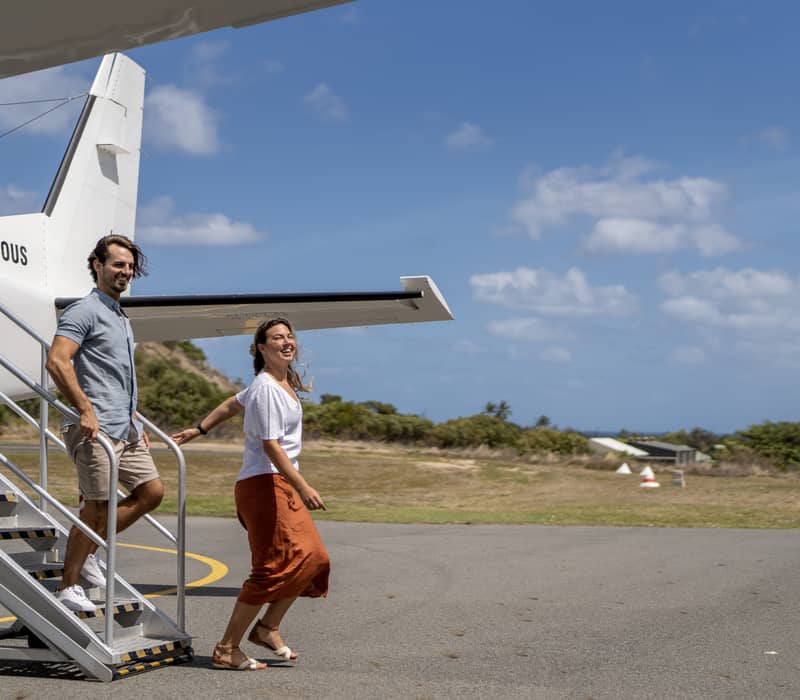 A smiling man and woman walking down the stairs of a small white airplane on a sunny day at an island airport.