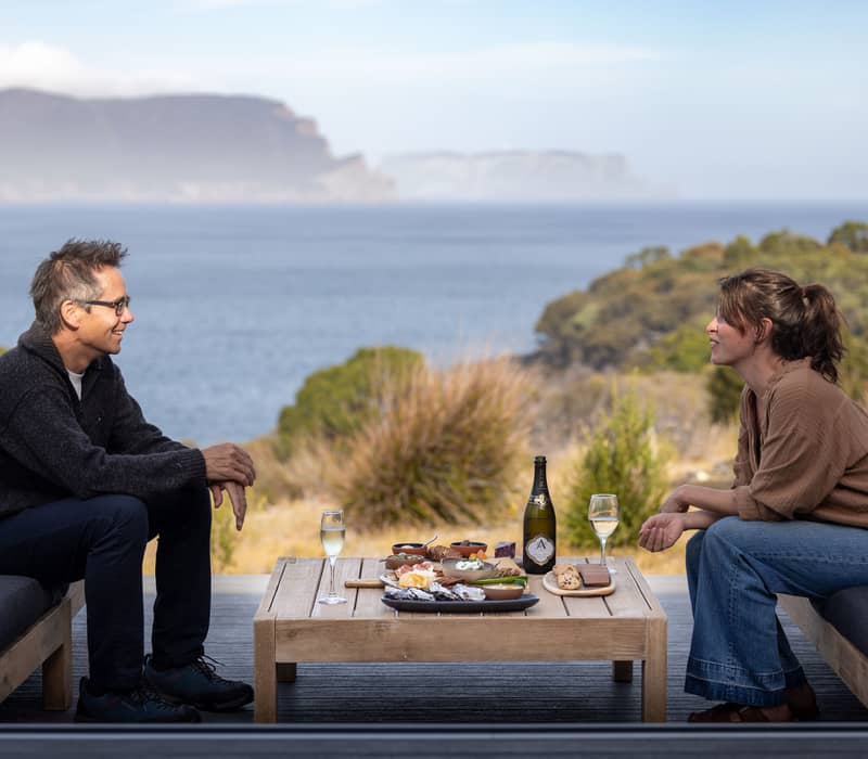 A man and woman sitting on outdoor sofas sharing oysters and wine with a mountain backdrop.
