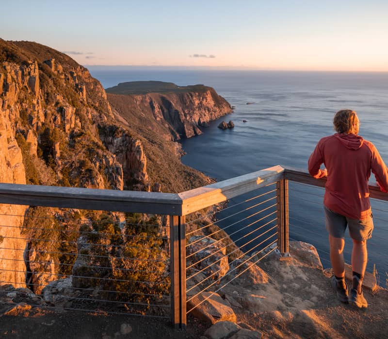 Man standing at a wooden lookout railing overlooking coastal cliffs during a golden sunset.
