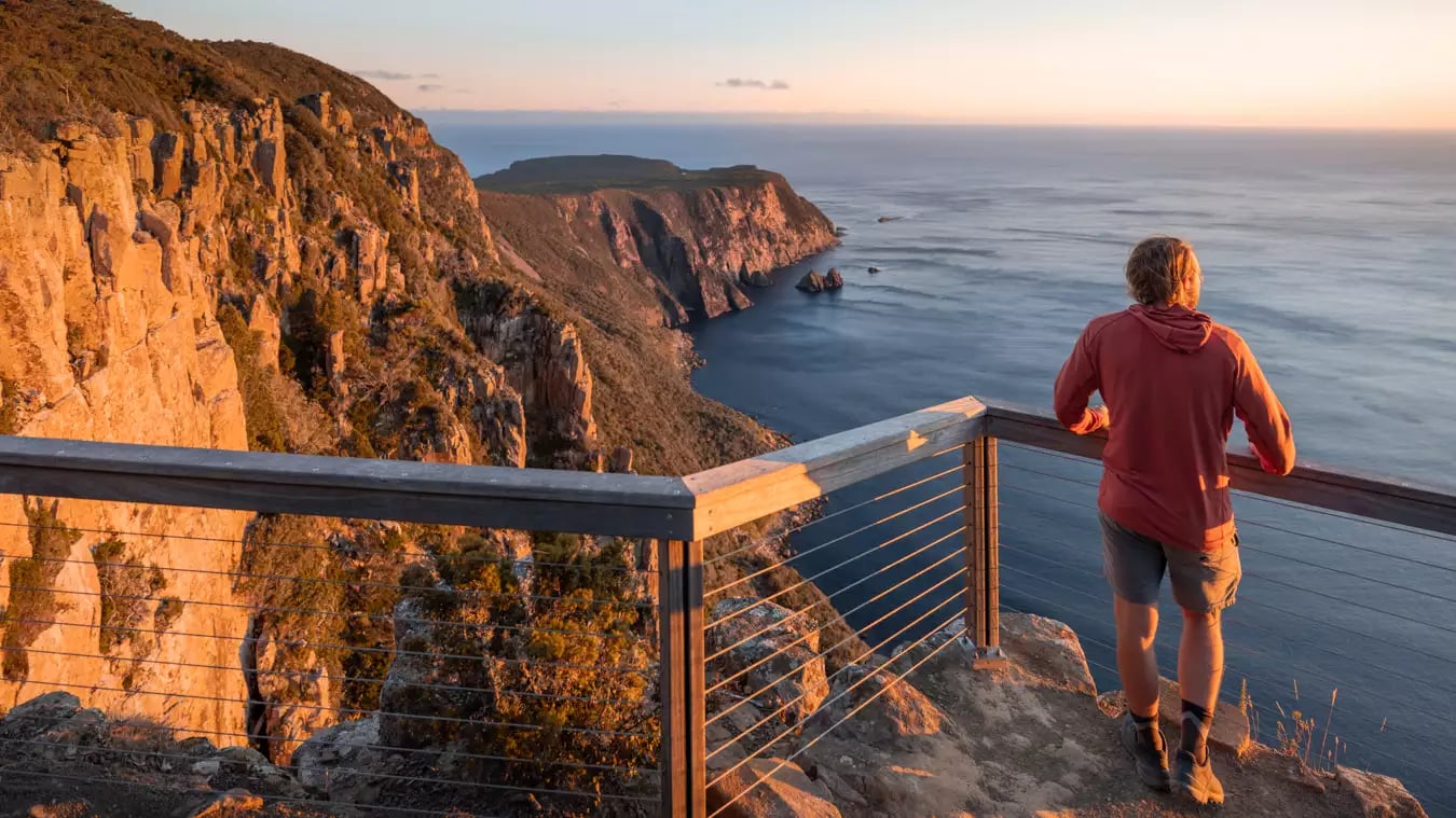 Man standing at a wooden lookout railing overlooking coastal cliffs during a golden sunset.