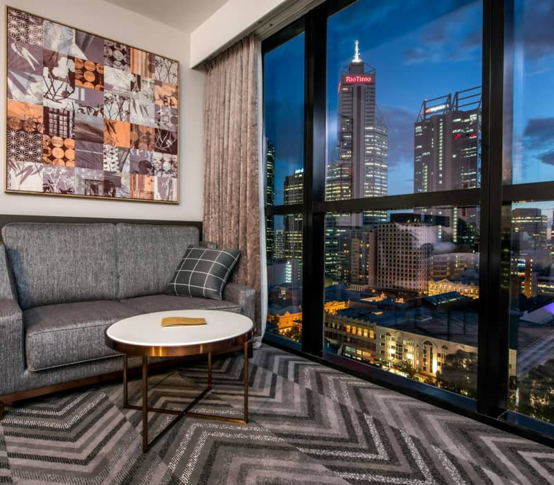 Contemporary hotel bedroom with a king bed, chevron-patterned carpet, and large windows overlooking Perth city.