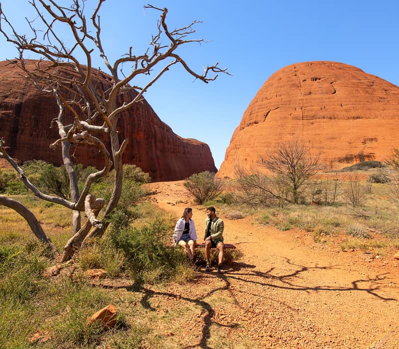 Two people rest on a bench in the sun-drenched desert with the massive, rounded red rock domes of Kata Tjuta rising behind them.