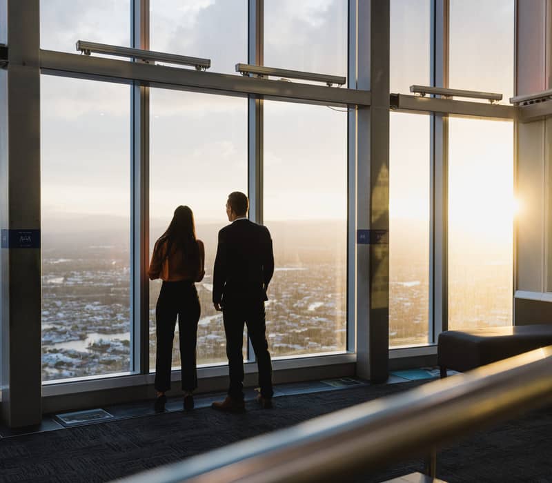 Silhouette of a man and woman looking out of floor-to-ceiling windows at a city sunset.
