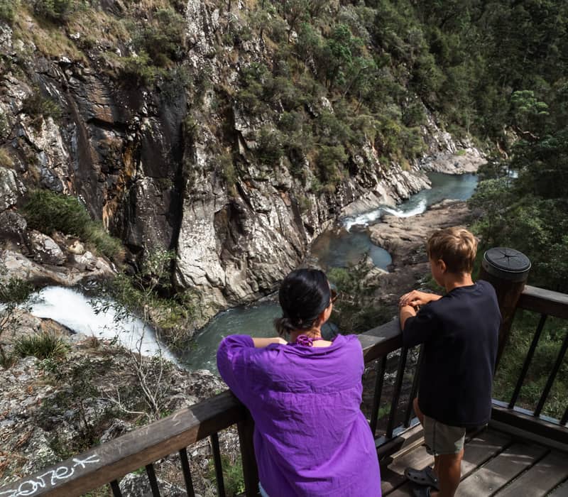 A woman and child leaning on a wooden railing looking down at a waterfall and rock pools in a canyon.