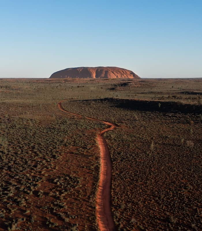 A high-angle drone shot captures a single orange-red track curving through a sea of green and brown desert bushes with Uluru on the horizon.