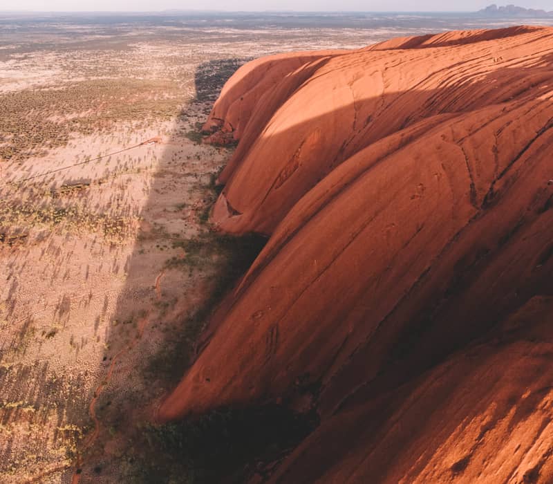 A wide drone perspective of the red monolith of Uluru rising from a sea of green desert scrub under a clear morning sky.