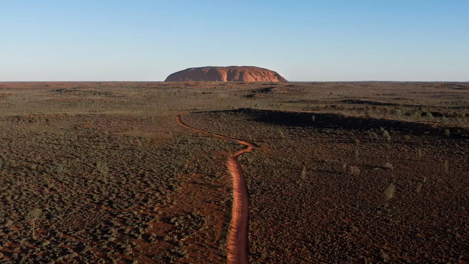 A high-angle drone shot captures a single orange-red track curving through a sea of green and brown desert bushes with Uluru on the horizon.