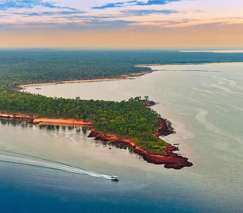 Aerial view of a boat on calm water near a forested coastal bay at sunset.